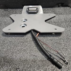 May include: A gray metal license plate bracket with an integrated LED light. The bracket has an X-shaped design with two circular holes and several mounting holes. A black wire harness and exposed wires extend from the bracket.