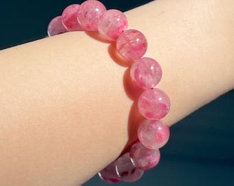 Puede incluir: Un brazalete de cuentas de piedras preciosas rosas. Las cuentas redondas y translúcidas presentan diferentes tonos de rosa. El brazalete se lleva en la muñeca, mostrando su color y diseño.