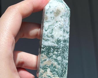 May include: A polished moss agate obelisk, featuring a pointed top and a rectangular base. The stone displays a mix of white, green, and tan colors, with intricate patterns. The obelisk is held in a hand.