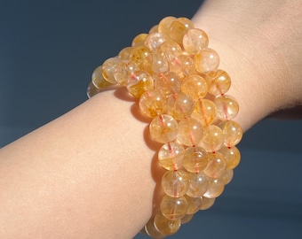 May include: A stack of three bracelets made of translucent, golden-yellow citrine beads. Each bead is round and has a slight variation in color, with some showing red inclusions. The bracelets are strung on a red cord.