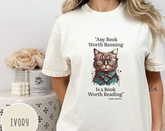Puede incluir: Una camiseta blanca con un gráfico de un gato con gafas y una pajarita. El texto en la camiseta dice "Any Book Worth Banning Is a Book Worth Reading" - Isaac Asimov.
