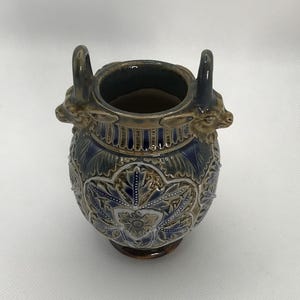May include: A small, blue and brown ceramic vase with two ram's head handles. The vase is decorated with intricate floral patterns and gold accents.