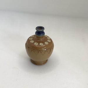 Puede incluir: Un pequeño jarrón de cerámica marrón con un diseño azul y blanco. El jarrón tiene un cuerpo redondo y un cuello estrecho.