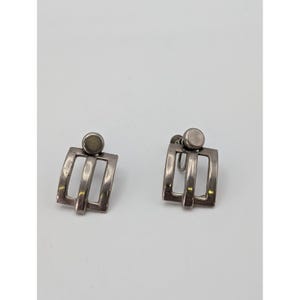 May include: A pair of silver-toned metal clip-on earrings. Each earring features a small circular top piece connected to a rectangular frame with three vertical bars. The design has a smooth, polished finish and a secure screw-back mechanism.