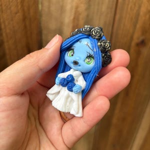 May include: A small, handcrafted figurine of a blue-skinned bride with blue hair and a black rose crown. She wears a white dress and holds blue roses. The figurine has green eyes and is held in a hand.