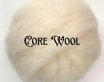 Pode incluir: Um close-up de uma bola de lã de núcleo esbranquiçada. A lã é fofa e texturizada, com as palavras "CORE WOOL" impressas em script preto no centro. O fundo é branco liso.