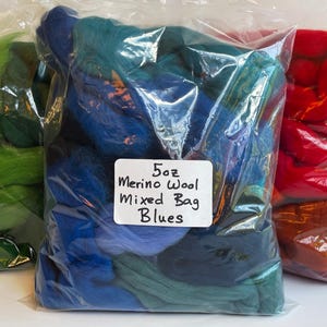May include: Three clear plastic bags filled with loose merino wool roving. The center bag contains various shades of blue wool and a white label reading 5oz Merino Wool Mixed Bag Blues. The left bag contains green wool, and the right bag contains red wool.