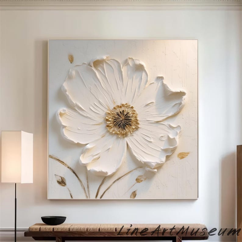 May include: A large, square, white floral art piece with a textured, raised design. The flower has a gold center and is set against a white background. The artwork is framed and hangs on a wall.