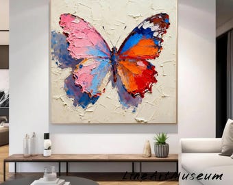May include: A large, textured painting of a butterfly with pink, blue, orange, and red wings. The artwork is set against a cream-colored background and framed in a light wood. The painting is displayed in a modern living space.