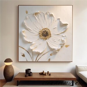 May include: A large, textured white flower art piece with a gold center and a brown frame. The artwork is mounted on a white background. The flower petals have a 3D effect. The artwork is a decorative piece.