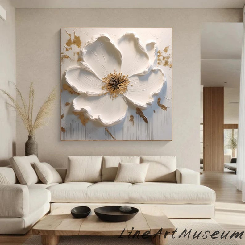 May include: A large, square white floral art piece with a textured, three-dimensional design. The flower has a golden center and gold accents on a white background. The artwork is displayed above a white sofa in a living room setting.