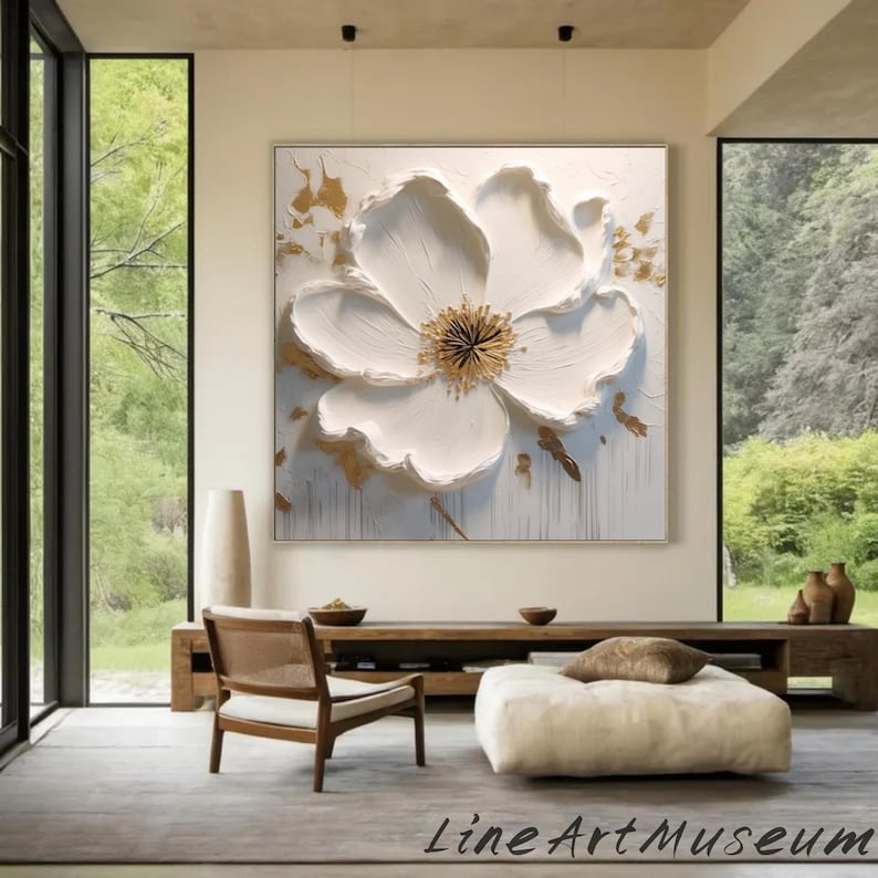 May include: Large white floral art piece with a textured, three-dimensional design. The flower has a golden center and gold accents on a white background. The artwork is framed and hangs on a wall in a modern living space.