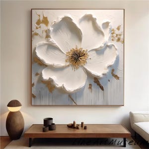 May include: Large, textured white flower art piece with gold accents. The artwork features a prominent, three-dimensional flower with a golden center, set against a white background. The piece is framed in a simple, dark wood frame, suitable for home decor.