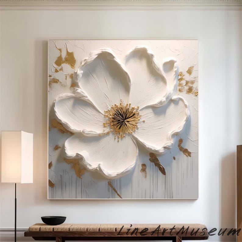 May include: Large, square white floral art piece with a textured, raised design. The flower has a gold center and gold accents on a white background. The artwork is framed and hangs on a white wall.