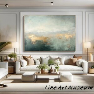Op de afbeelding: Groot abstract schilderij met goud- en blauwgroene tinten. Het kunstwerk hangt boven een witte bank met sierkussens. De kamer is ingericht in neutrale kleuren en bevat de tekst "Line Art Museum".