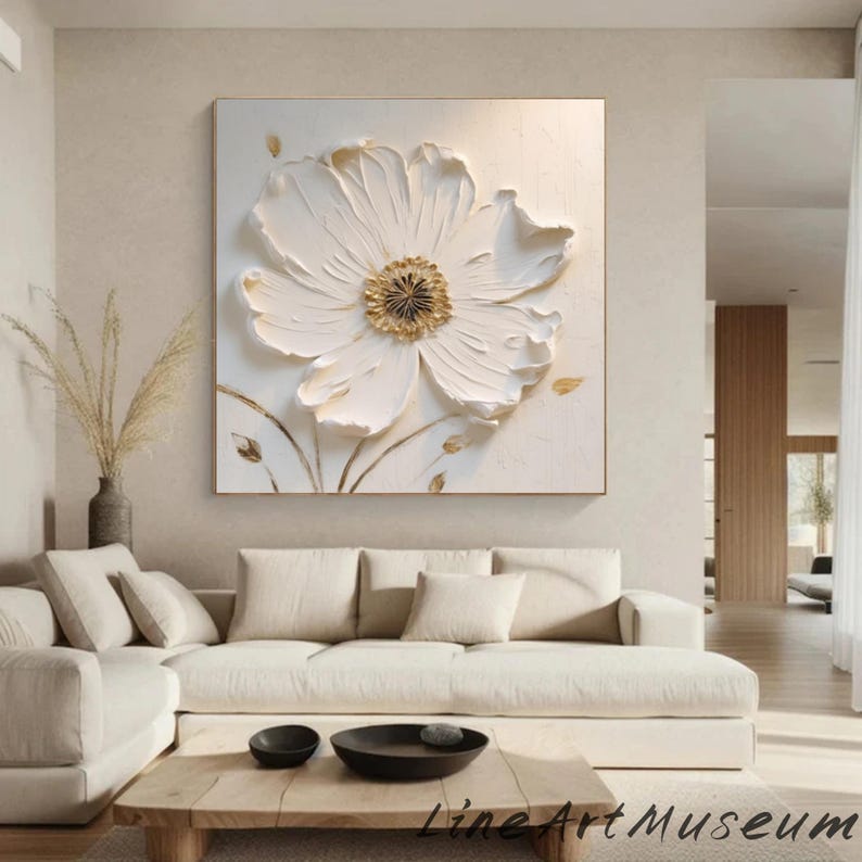 May include: A large, square, white floral painting with a textured, three-dimensional effect. The flower has a golden center and is set against a neutral background. The artwork is framed in a gold-colored frame.