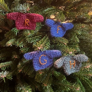May include: Four miniature knitted sweaters with different colors and letters embroidered on them. The sweaters are hanging on a green Christmas tree.