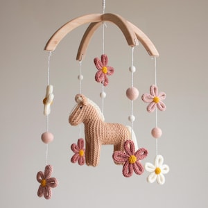 May include: A hanging mobile featuring a central knitted tan horse figure surrounded by crocheted flowers in shades of dusty rose and cream. The elements are suspended from a curved light wood frame by white strings accented with small felt balls.