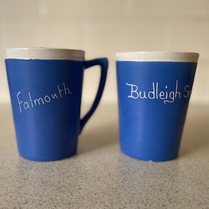 Może przedstawiać: Dwa niebieskie ceramiczne kubki z białymi obwódkami. Jeden kubek ma napis "Falmouth", a drugi "Budleigh Salterton".