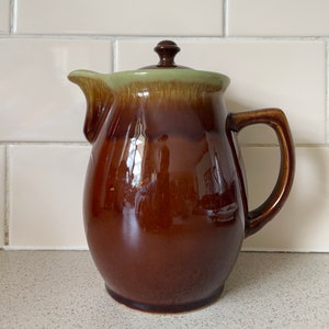 May include: A brown and green glazed ceramic coffee pot with a lid. The pot has a rounded body and a handle.