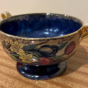 Op de afbeelding: Een blauwe en witte keramische kom met gouden handvatten en bloemmotieven. De kom heeft een gegolfde rand en een voet.