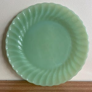 May include: A round, light green jadeite plate featuring a smooth, flat centre and a decorative, swirling, fluted edge design. The translucent glass material has a soft, pale mint hue with a glossy finish throughout the entire surface.