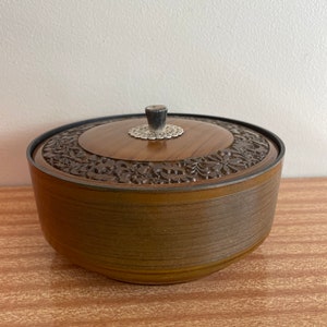 May include: A round wooden box with a carved floral design and a black knob on the lid. The box has a brown wood grain finish and a black rim around the top.