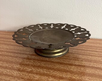 May include: A small, round, brass trinket dish with a decorative, scalloped edge and a raised, circular base. The dish has a delicate, intricate design of small, repeating shapes.
