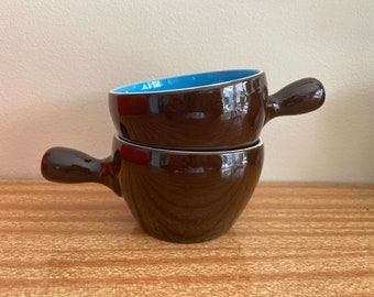 May include: Two brown ceramic bowls with blue rims and handles. The bowls are stacked on top of each other.
