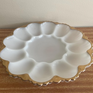 May include: A white milk glass egg plate featuring twelve oval indentations arranged in a circular pattern around a smooth center. The scalloped edge of the plate is finished with a metallic gold trim that follows the curved contour of the rim.
