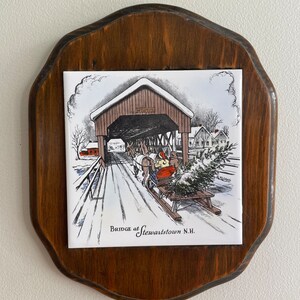 May include: A wooden plaque with a white tile featuring a black and white illustration of a covered bridge in a snowy setting. The text "Bridge at Stewartstown N.H." is printed below the image.