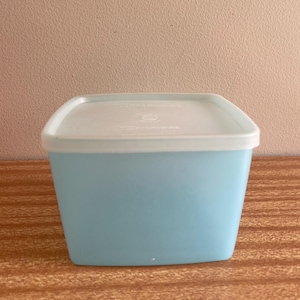 Pode incluir: Um recipiente quadrado de plástico azul claro com uma tampa branca. O recipiente tem um fundo ligeiramente arredondado e a tampa tem um centro ligeiramente elevado.