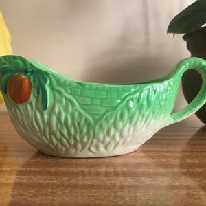 May include: A green ceramic gravy boat with a white interior and a handle. The boat is decorated with a leafy design and a single orange fruit.