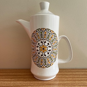 May include: White ceramic coffee pot with a brown, green, and yellow geometric design. The pot has a lid and a handle.