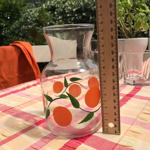 May include: A clear glass pitcher with an orange and green citrus fruit design. The pitcher is approximately 7 inches tall. It sits on a checkered tablecloth. A ruler is next to the pitcher.