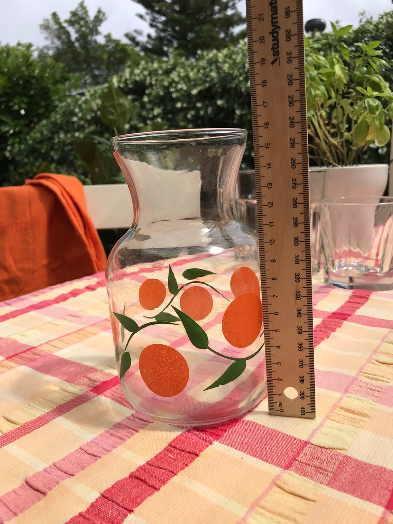May include: A clear glass pitcher with an orange and green citrus fruit design. The pitcher is approximately 7 inches tall. It sits on a checkered tablecloth. A ruler is next to the pitcher.