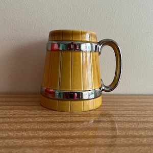 May include: A yellow ceramic mug with a silver rim and handle, shaped like a barrel.