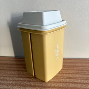 May include: A mustard yellow plastic storage container with a white lid. The container has a rectangular shape with rounded corners and a vertical slit along the side. A small white floral graphic is printed on the front of the yellow body.