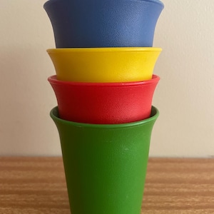 May include: A stack of four brightly coloured plastic cups. The cups are in primary colours: green, red, yellow, and blue. The cups are stacked, with the green cup at the bottom and the blue cup at the top. The cups have a textured surface.
