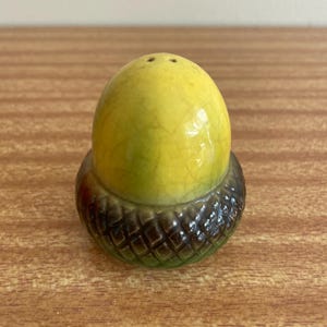 Op de afbeelding: Een keramische zoutstrooier in de vorm van een eikel. De bovenkant is geel met een gebarsten glazuur en de basis is bruin met een textuurpatroon. De strooier heeft drie kleine gaatjes aan de bovenkant.