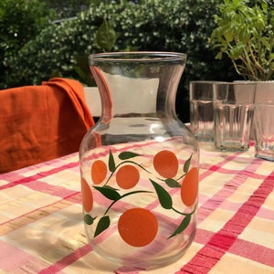 May include: A clear glass pitcher with a wide base and a flared top. The pitcher is decorated with orange fruit and green leaves. It sits on a checkered tablecloth with other glasses in the background.