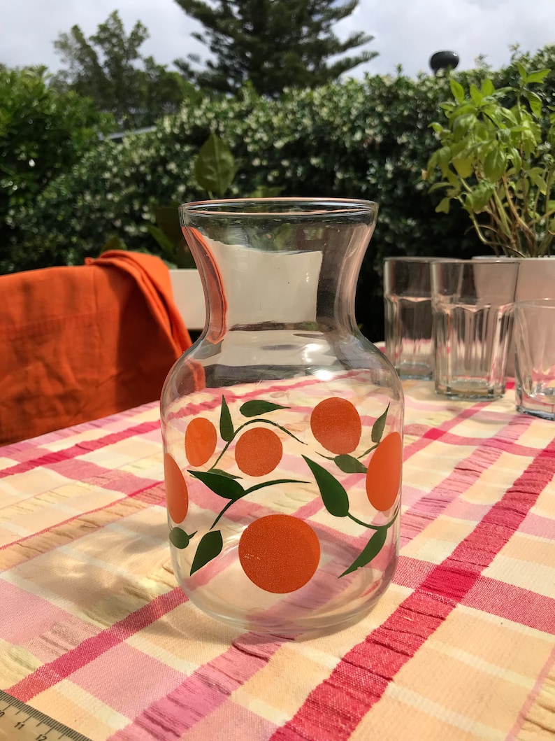 May include: A clear glass pitcher with a wide base and a flared top. The pitcher is decorated with orange fruit and green leaves. It sits on a checkered tablecloth with other glasses in the background.