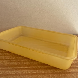 Könnte beinhalten: Ein rechteckiger, durchscheinender gelber Kunststoffbehälter. Der Behälter hat eine geringe Tiefe und abgerundete Ecken. Er steht auf einer Holzoberfläche.