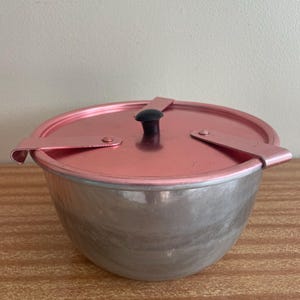 Op de afbeelding: Een vintage popcornpoffer met een roségouden deksel en rand, en een zilverkleurige kom. Het deksel heeft een zwarte knop en twee handvatten. De poffer staat op een houten oppervlak.