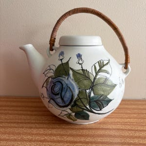 May include: A round white ceramic teapot featuring a hand-painted blue rose and green leaves. The teapot has a short spout, a matching lid, and a curved, textured brown rattan handle attached to two side loops on the body of the vessel.