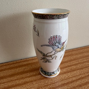 May include: A white porcelain vase featuring a colourful hummingbird perched on a branch. The vase has a flared rim and a circular base, both decorated with a dark, mottled pattern and gold trim. The body displays a delicate floral illustration.