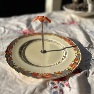 May include: A vintage cake stand with a white ceramic plate and a decorative orange, yellow, and blue floral design. The stand has a metal rod with a pink plastic handle.