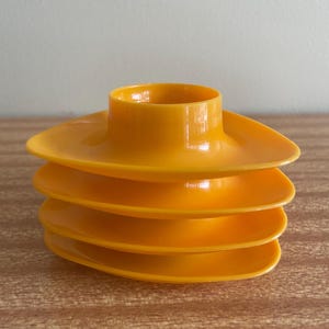 May include: A stack of four bright orange plastic egg cups. Each egg cup features a rounded triangular base with a central circular holder designed to support an egg. The smooth, glossy finish of the plastic is consistent across all four pieces.