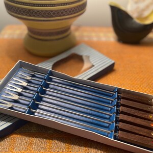 May include: A vintage fondue set with stainless steel forks and wooden handles with coloured accents. The set is in a white box with blue dividers and a striped lid. Other decorative items are in the background.