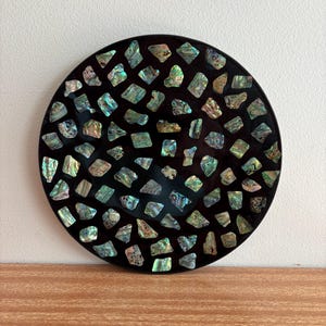 May include: A round, black decorative plate inlaid with iridescent, irregularly shaped pieces. The plate's surface is covered with a mosaic of shimmering, colorful fragments, creating a unique and eye-catching design. The plate is displayed on a wooden surface.